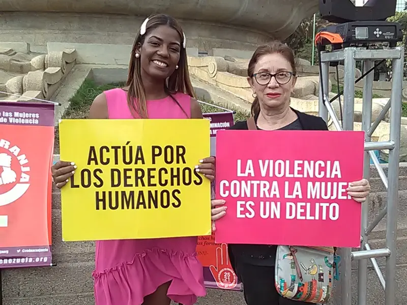 Dos mujeres sostienen carteles que dicen «Actúa por los derechos humanos» y «La violencia contra la mujer es un delito» durante una jornada al aire libre en Caracas